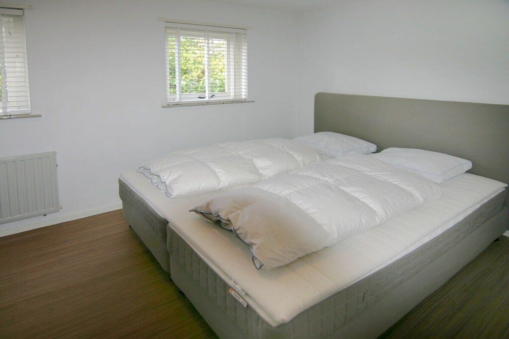 Een kamer met een enkel bed, een grijs hoofdeinde, wit beddengoed en een raam met zonwering.