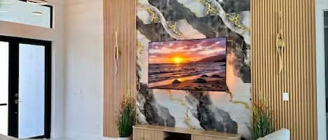 A modern living room with a large sofa, a ceiling fan, and a wall-mounted TV displaying a sunset scene.