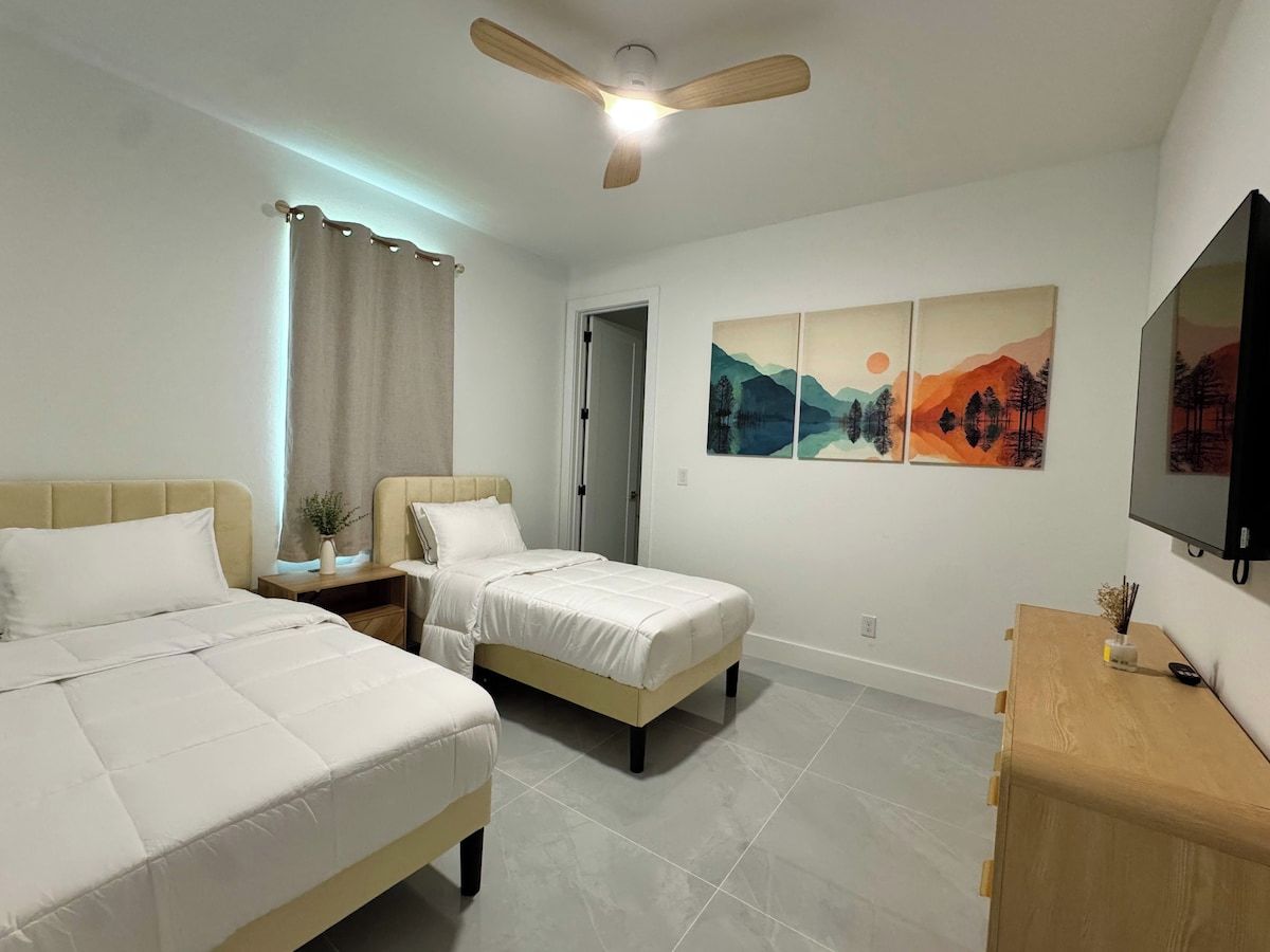 Habitación de hotel con dos camas, ventilador de techo, televisor y obras de arte en la pared.