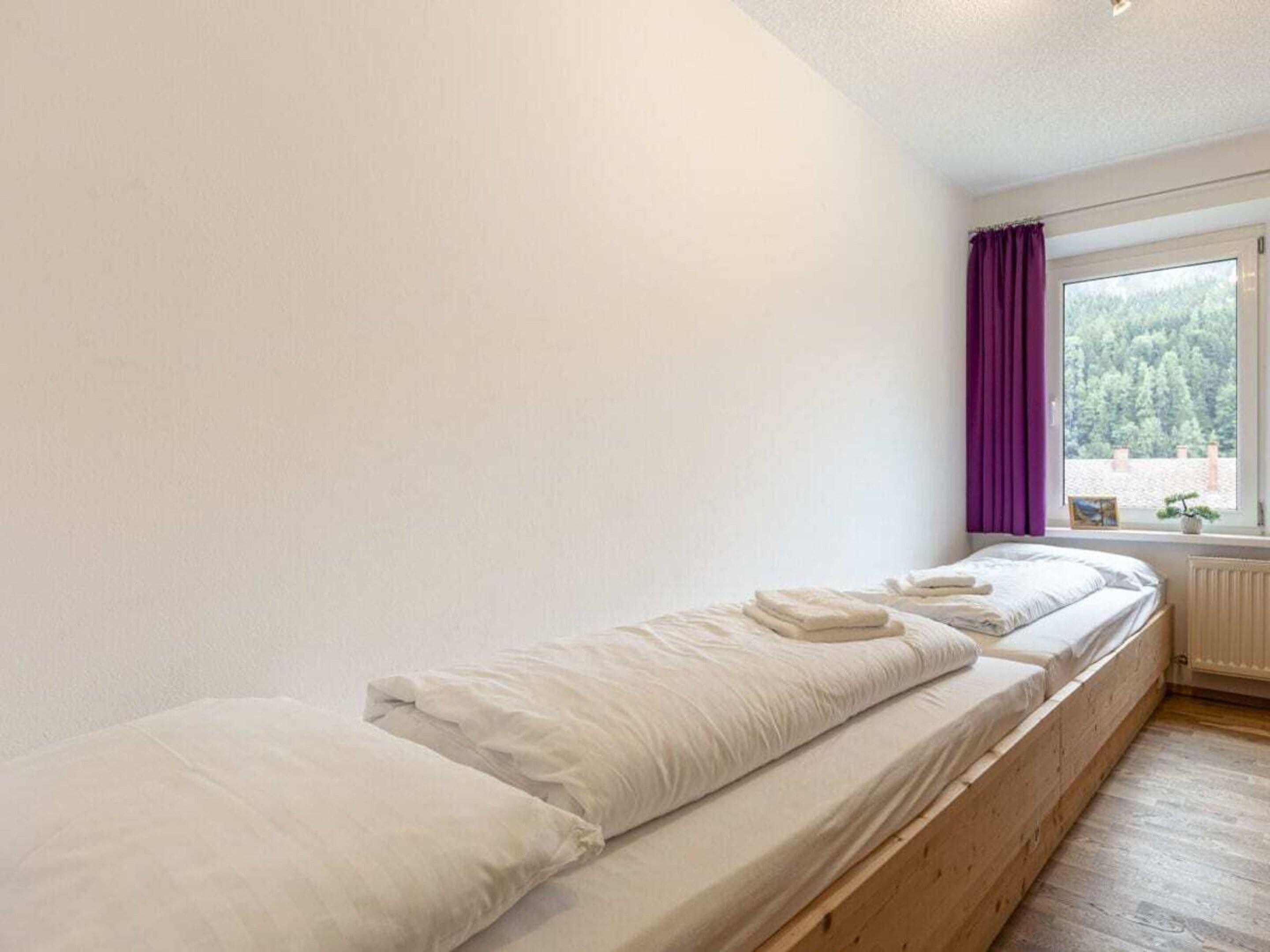 Een eenpersoonsbed met wit beddengoed in een kamer met witte muren en een raam met paarse gordijnen.