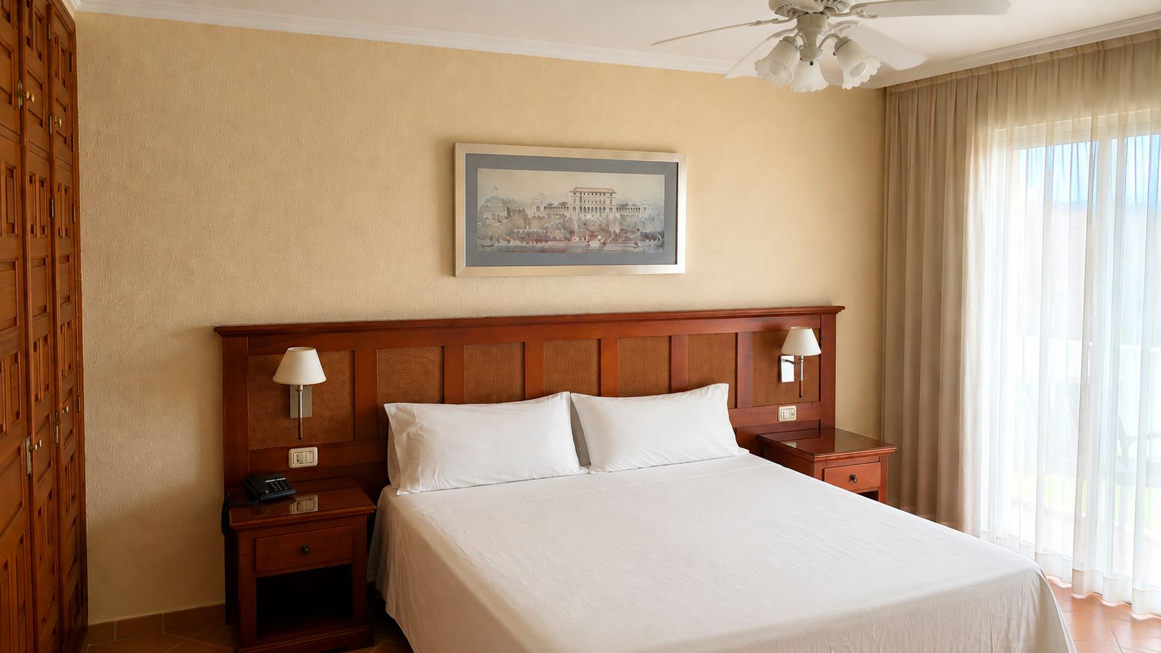 Habitación de hotel con una cama grande, cabecero de madera, dos mesitas de noche con lámparas, una mesita auxiliar y un cuadro en la pared.