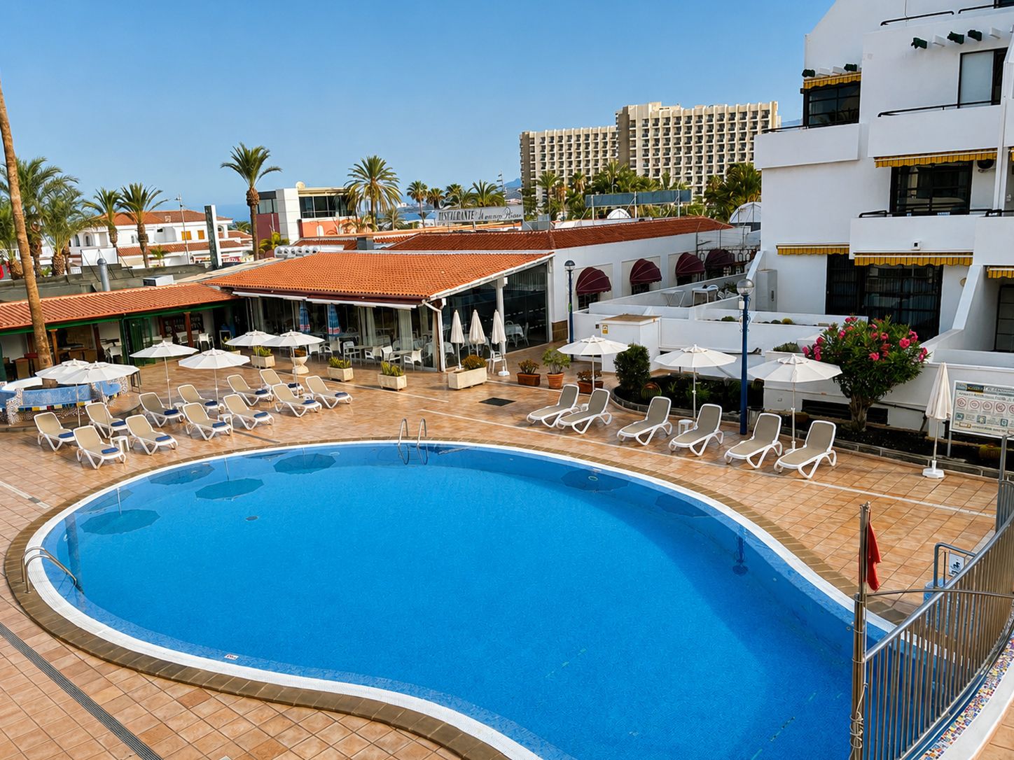 A swimming pool with lounge chairs and umbrellas, surrounded by buildings and palm trees.