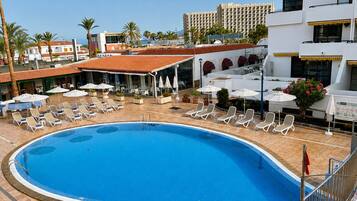 A swimming pool with lounge chairs and umbrellas, surrounded by buildings and palm trees.