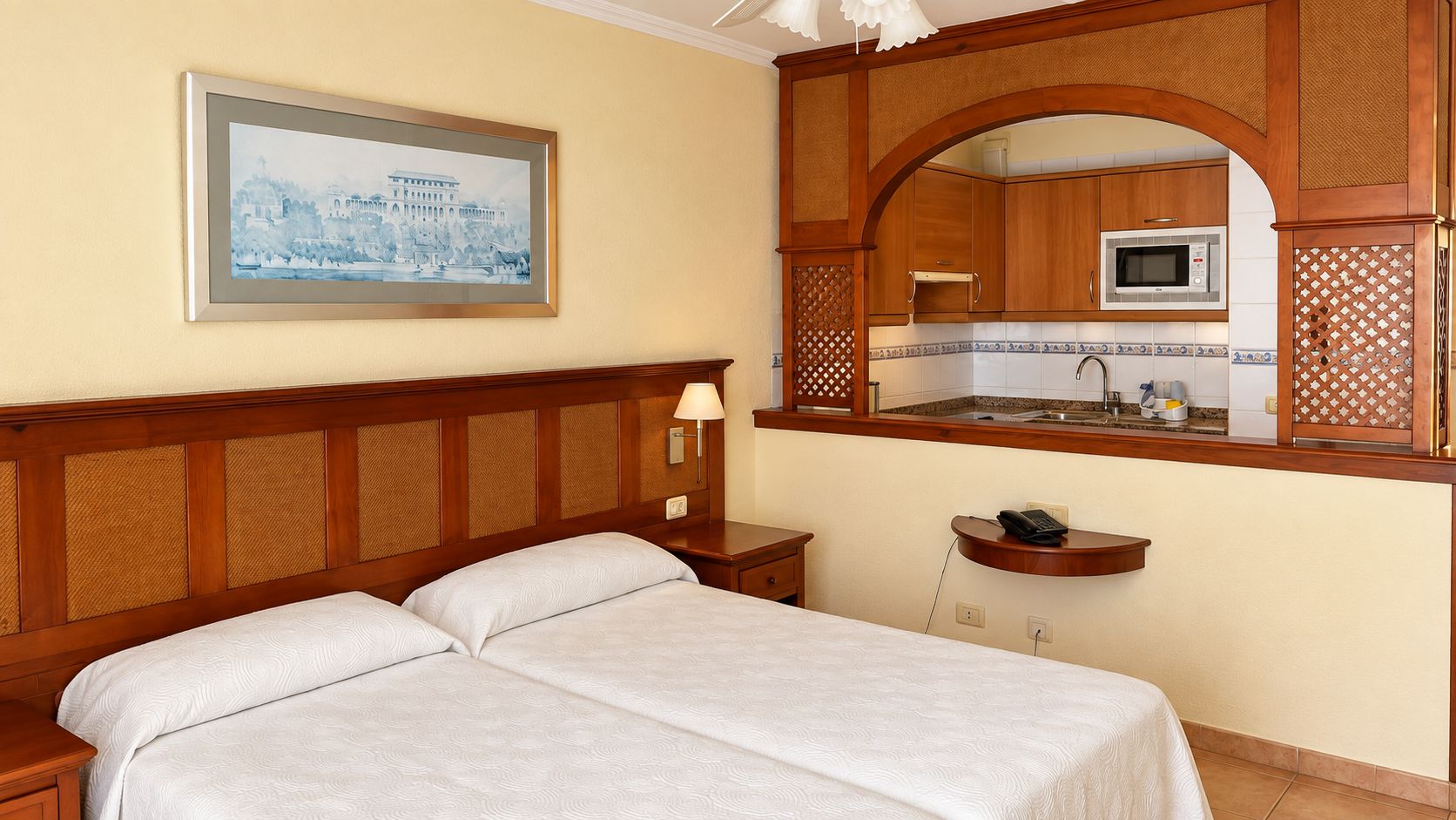 Een hotelkamer met een bed, nachtkastje, lamp en uitzicht op de keuken door een raam met houten kader.