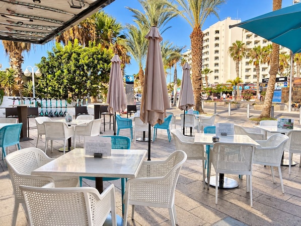 An outdoor cafe area with tables and chairs, umbrellas, and palm trees.