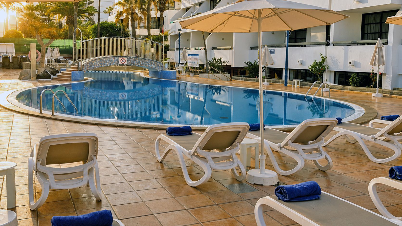 Piscina con sillones de descanso y sombrillas, rodeada de una terraza con baldosas y palmeras.