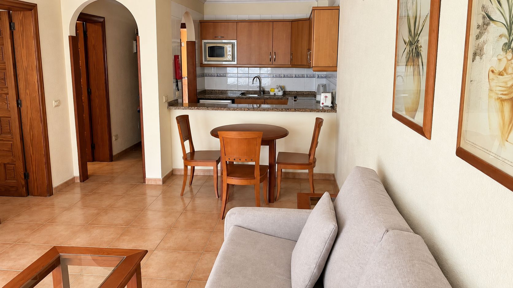 A living room with a sofa, a dining table with chairs, a kitchen area with wooden cabinets and a microwave, and a framed picture on the wall.