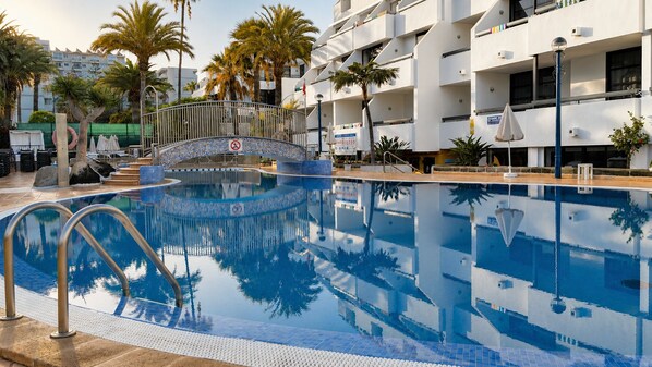 A swimming pool with a bridge, palm trees, and a building with balconies.