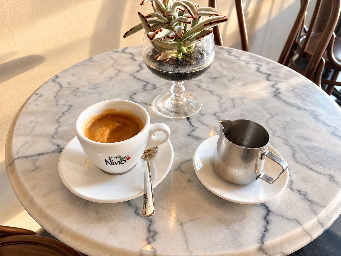 Une table ronde en marbre, sur laquelle se trouvent une tasse à café blanche, un pichet en métal et un verre contenant une plante.