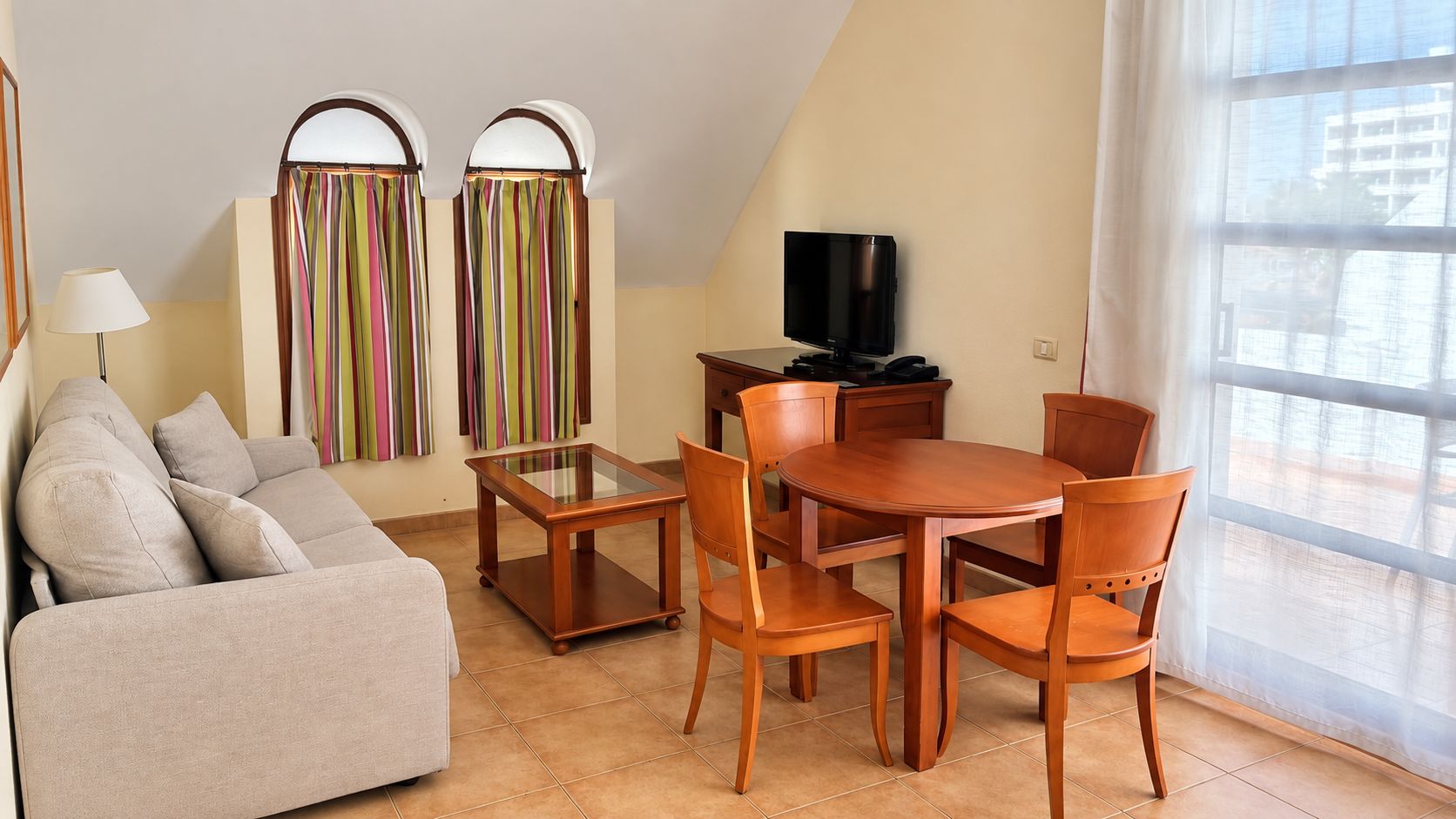 Una sala de estar moderna con un sofá, una mesa de centro de vidrio, sillas de madera, un televisor y un ventanal con cortinas.