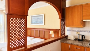 A hotel room with a wooden partition, a bed, a nightstand with a lamp, a kitchen area with a toaster and kettle, and a framed picture on the wall.