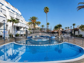 A swimming pool with a tiled deck, surrounded by palm trees and a white building with balconies.