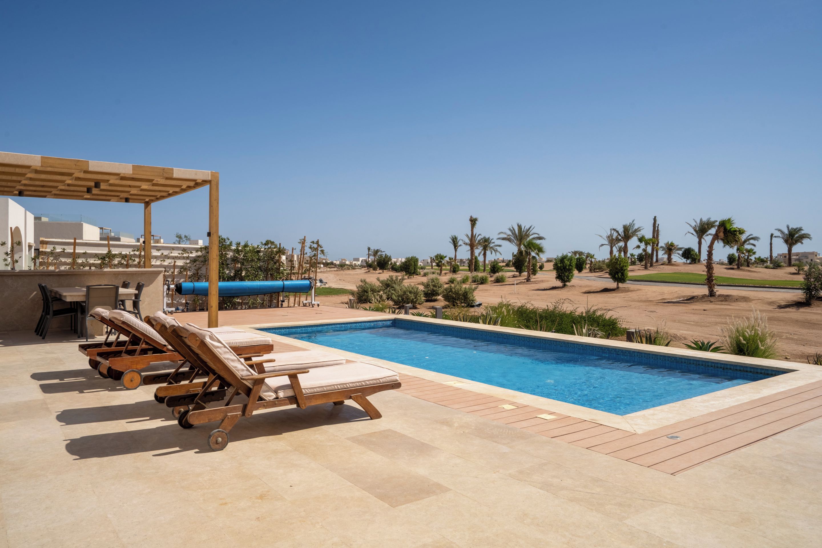A swimming pool with lounge chairs and a pergola, surrounded by palm trees and a clear sky.