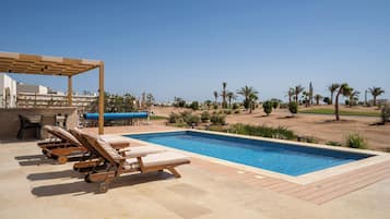 A swimming pool with lounge chairs and a pergola, surrounded by palm trees and a clear sky.