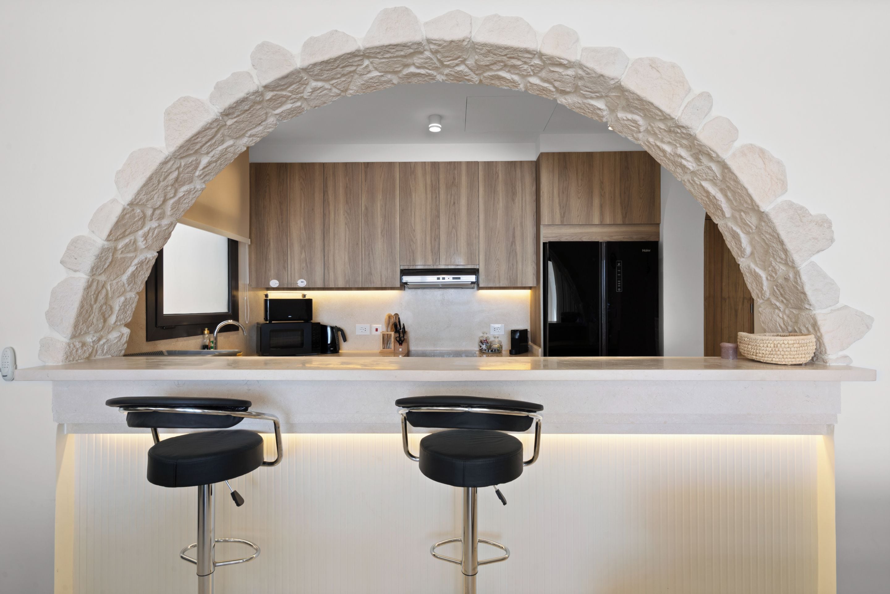 A modern kitchen with a stone arch, wooden cabinets, a black refrigerator, and two bar stools.