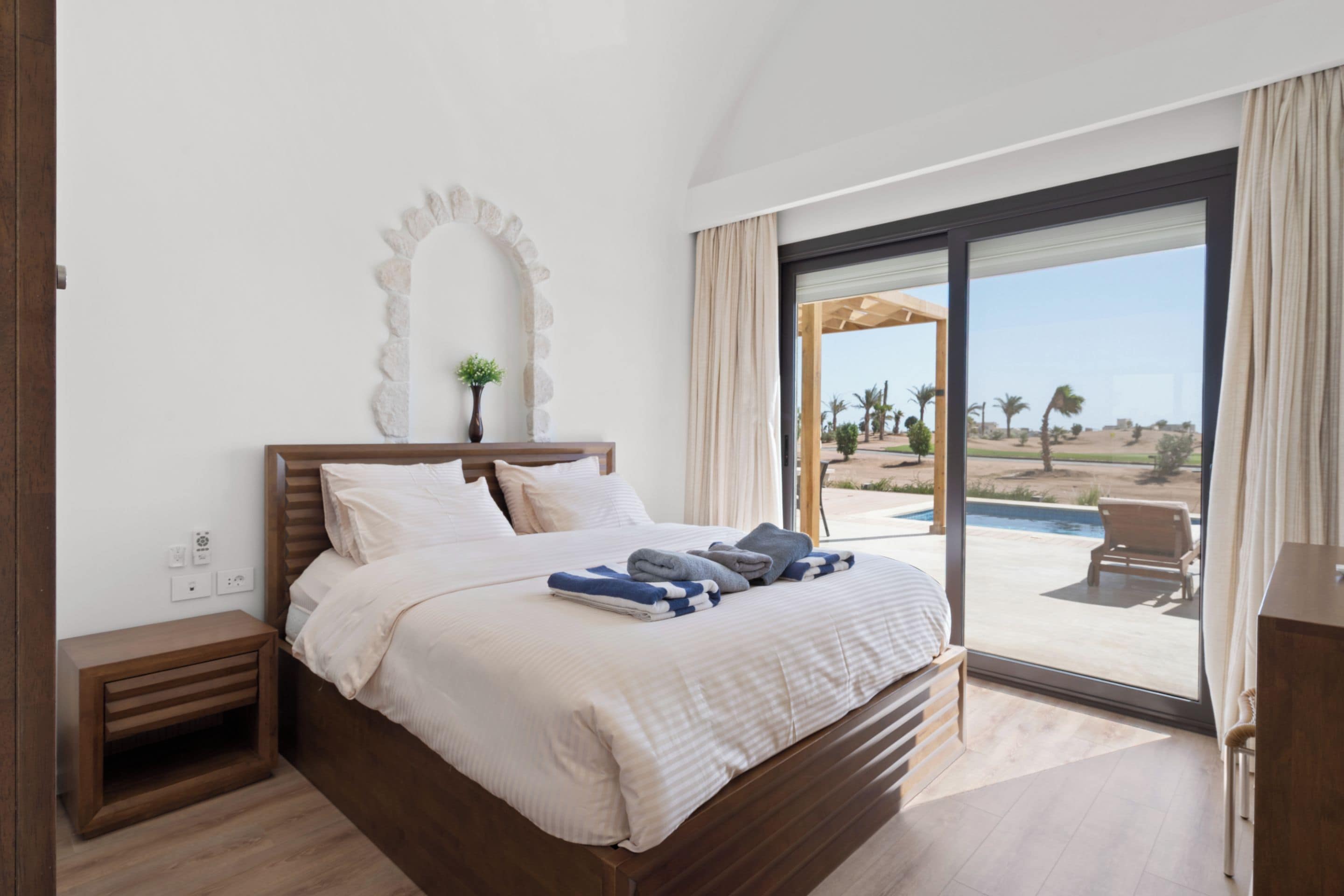 A bedroom with a large bed, a nightstand, a wall-mounted mirror, and a view of a pool and outdoor area.