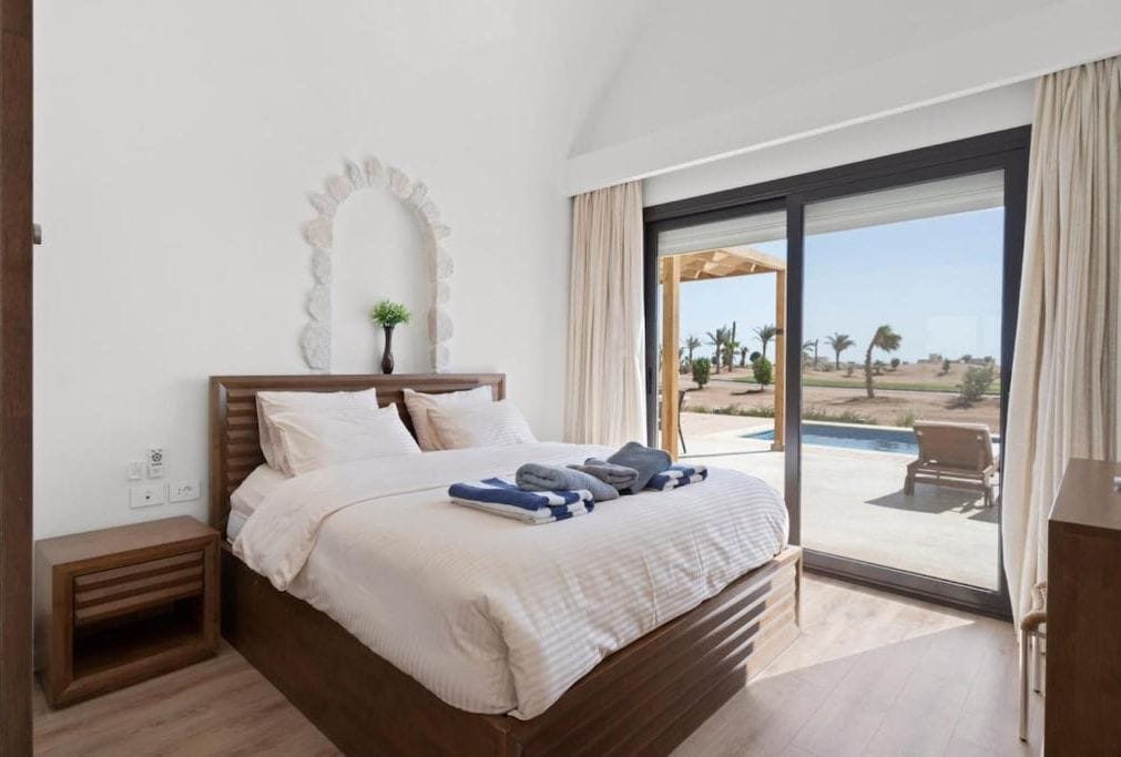 Ein Schlafzimmer mit einem großen Bett, einem Nachttisch, einem Spiegel und Blick auf Pool und Außenbereich.