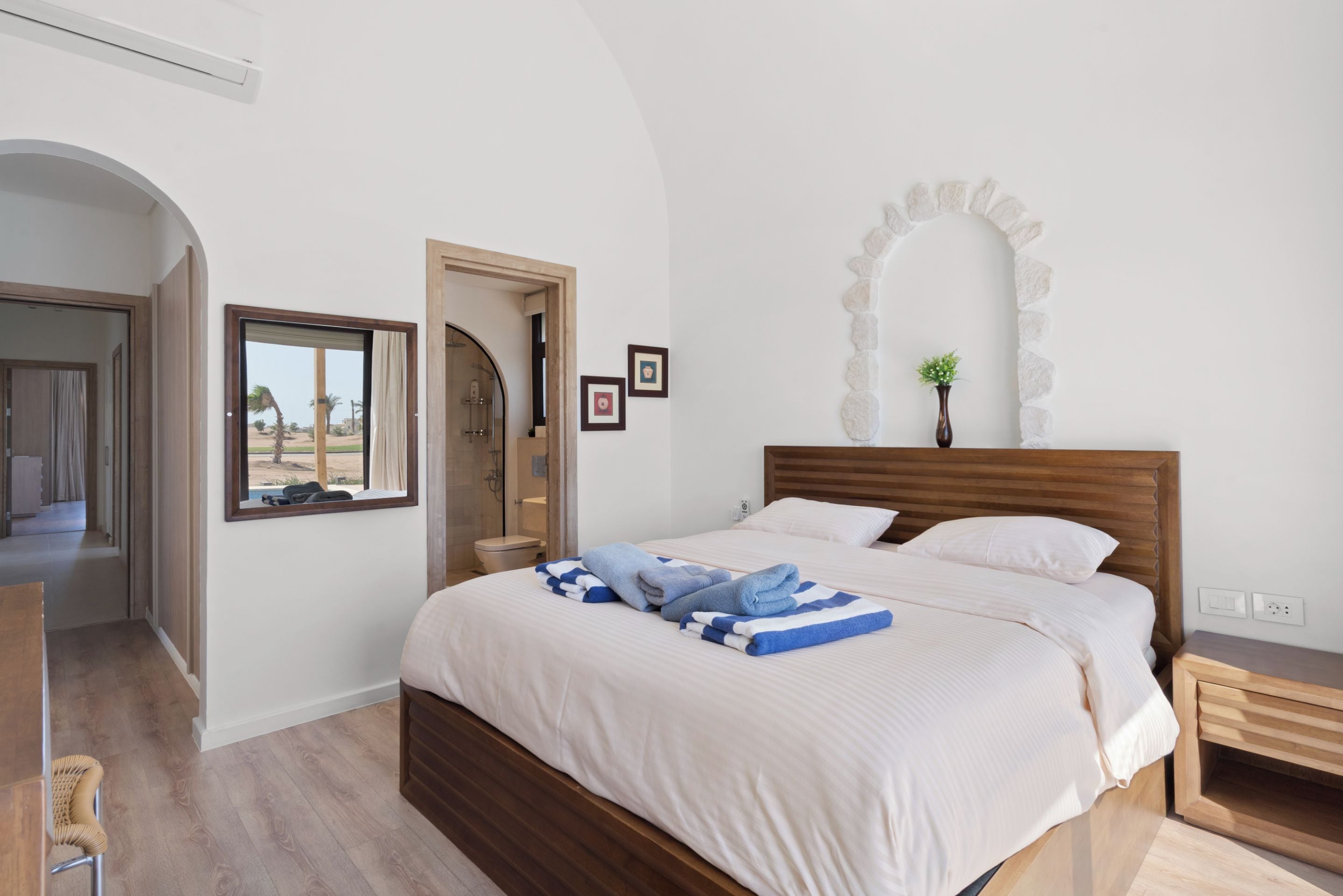 A bedroom with a large bed, a wooden headboard, a window with a view, a mirror, and a small plant on the wall.