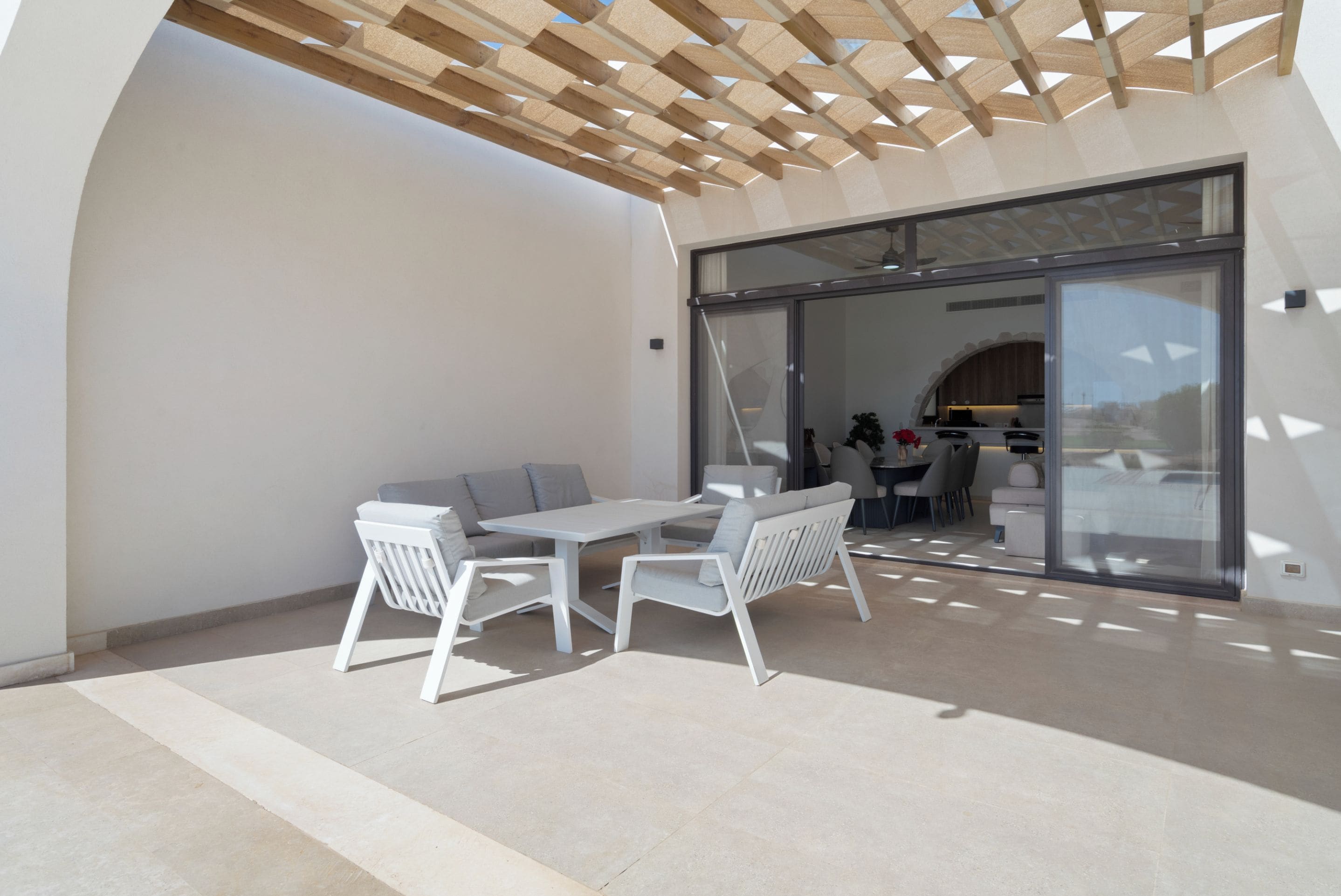 Une terrasse extérieure avec une table et des chaises blanches, une pergola en bois au-dessus, et une porte vitrée donnant sur un espace intérieur.