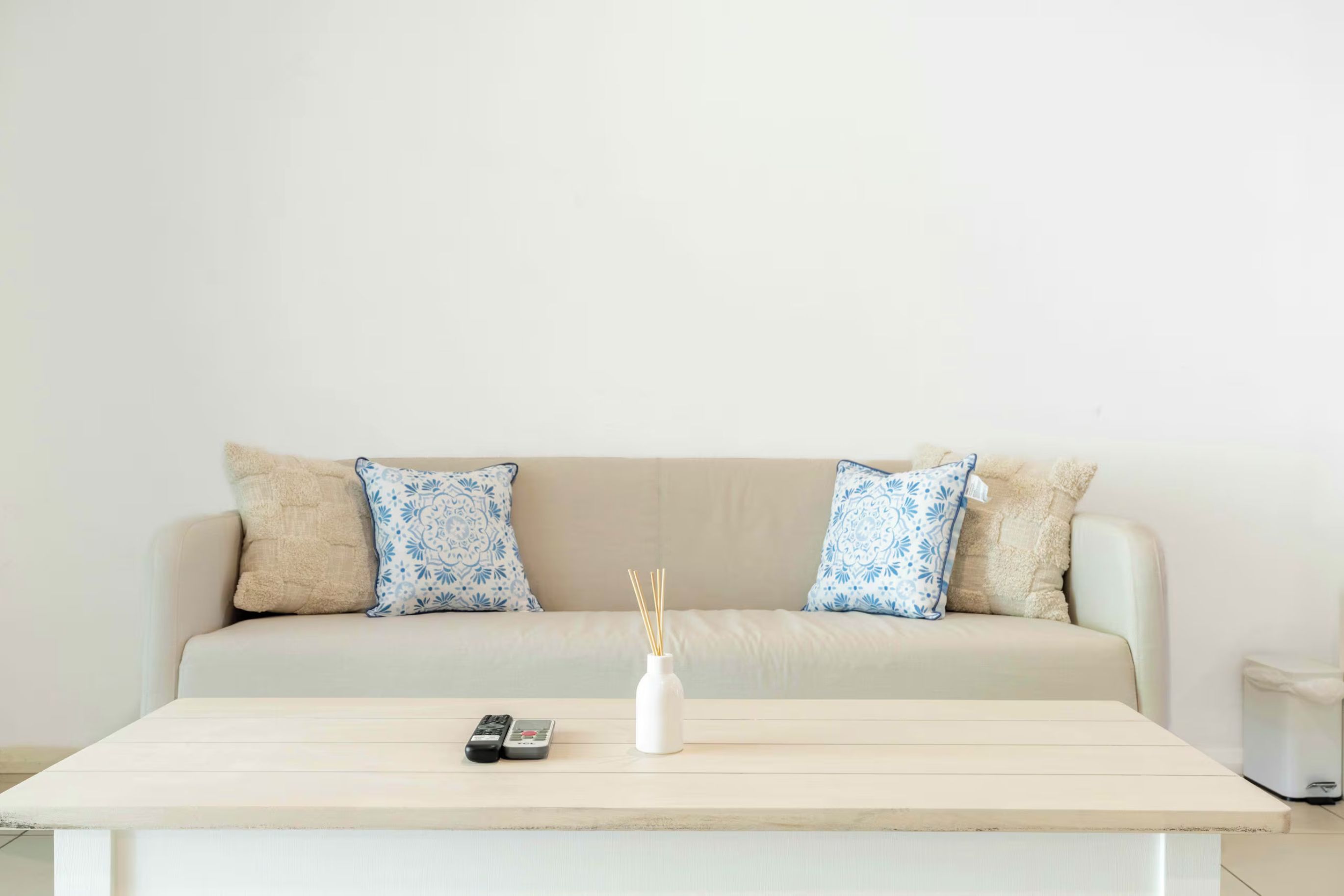 A neutral-colored sofa with two patterned pillows, a remote control, and a white vase with reed sticks on a light-colored table.