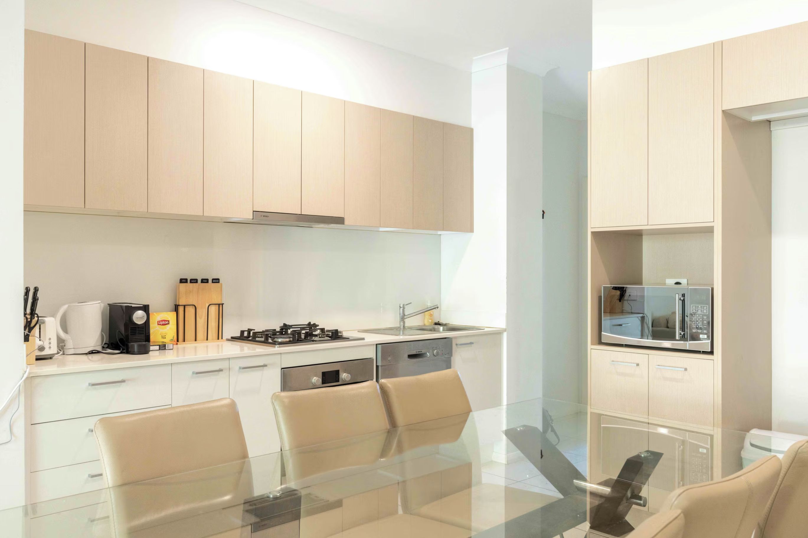 A modern kitchen with light wood cabinets, a gas stove, a dishwasher, and a dining area with a glass table and chairs.