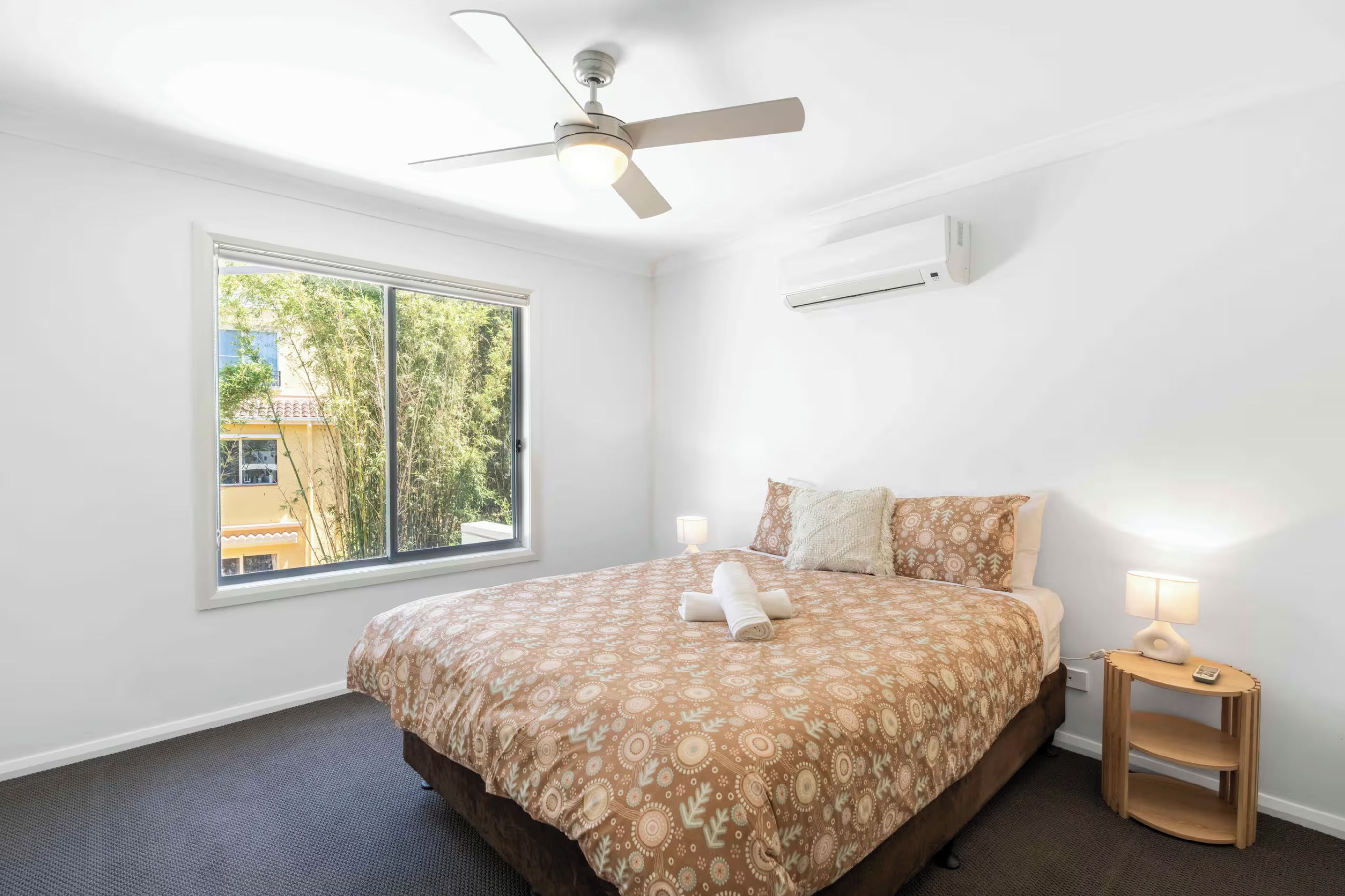A bedroom with a bed, a ceiling fan, a window, and a bedside table with a lamp.