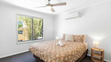 A bedroom with a bed, a ceiling fan, a window, and a bedside table with a lamp.