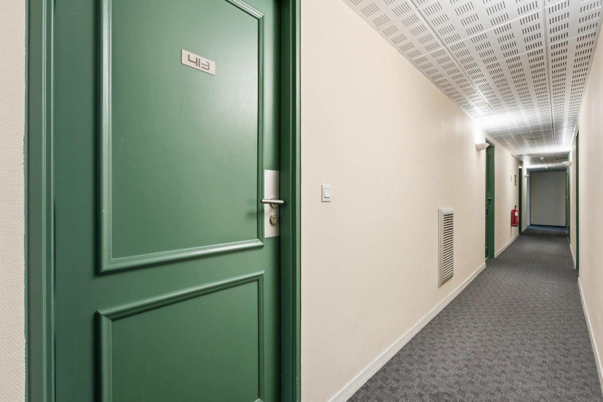 A hallway with green doors, a fire alarm, and a ceiling with a grid pattern.