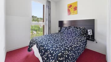 Une chambre à coucher avec un lit, une table de chevet, une lampe, une fenêtre avec des rideaux et un tableau au mur.