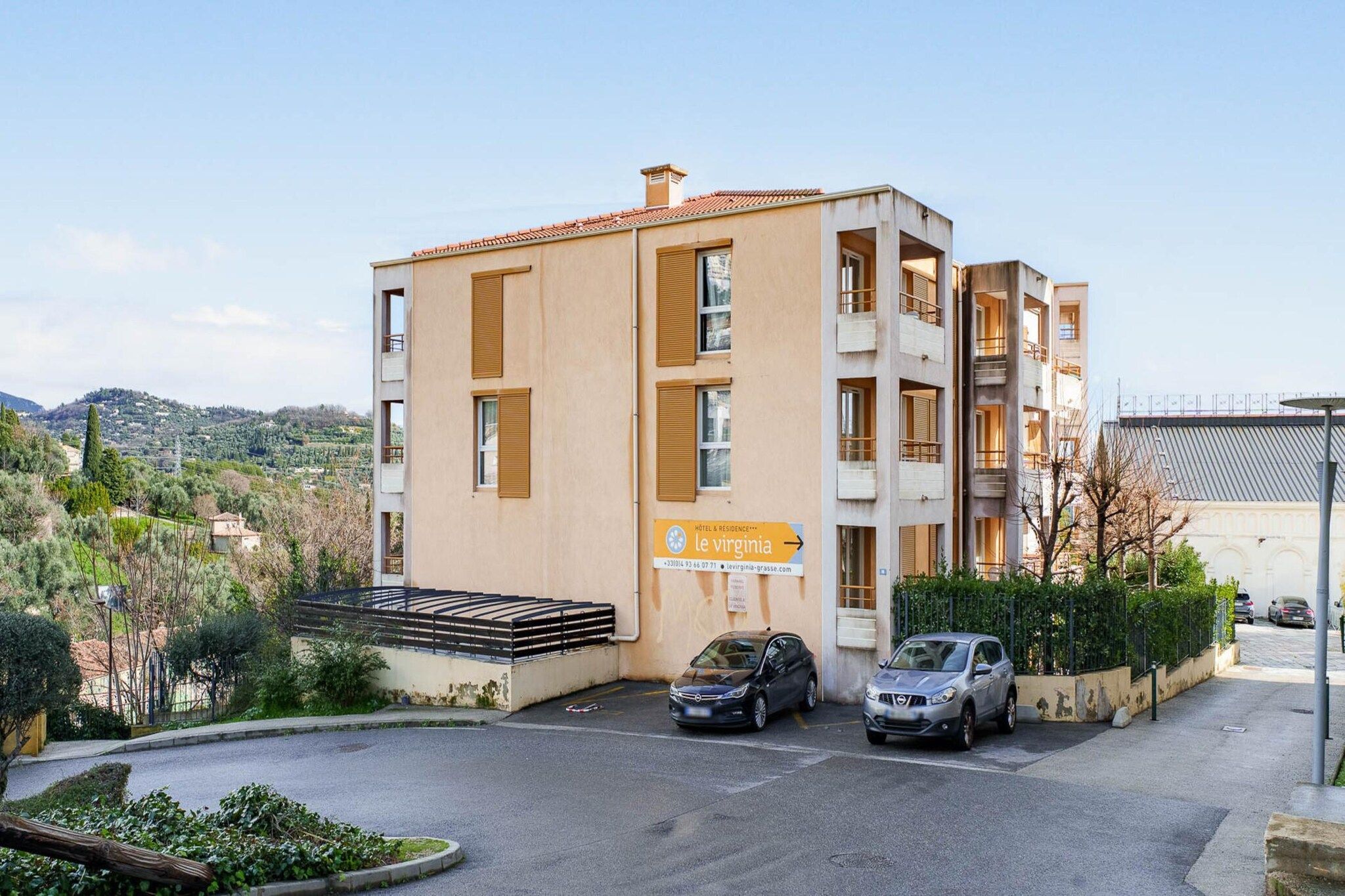 Un immeuble d’hôtel avec une enseigne indiquant « Hôtel et résidence Le Virginia ». Deux voitures sont garées devant, et l’immeuble compte plusieurs étages avec des balcons.