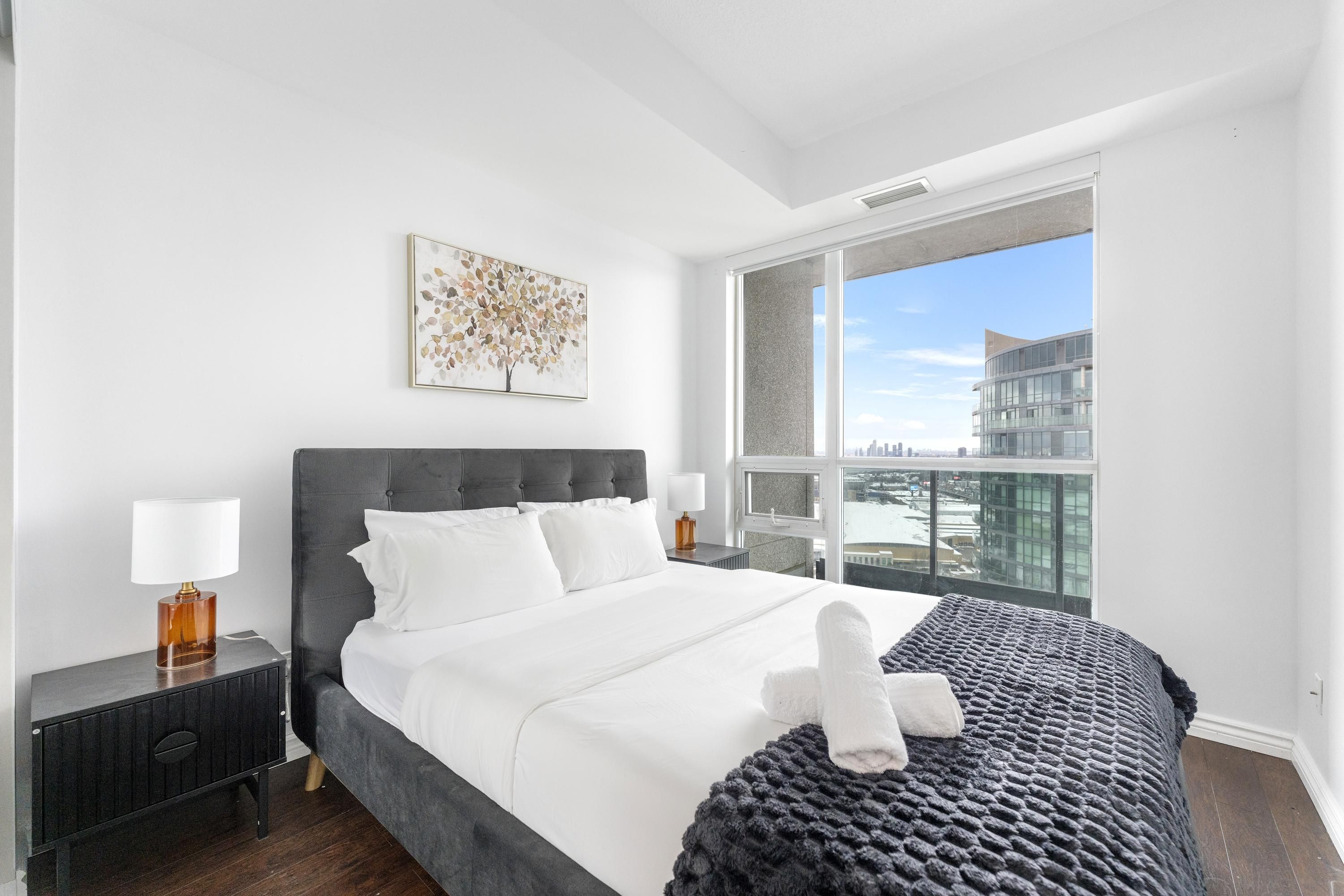 Un dormitorio moderno con una cama grande, una mesita de noche, una lámpara, una ventana con vistas a la ciudad y una obra de arte enmarcada en la pared.