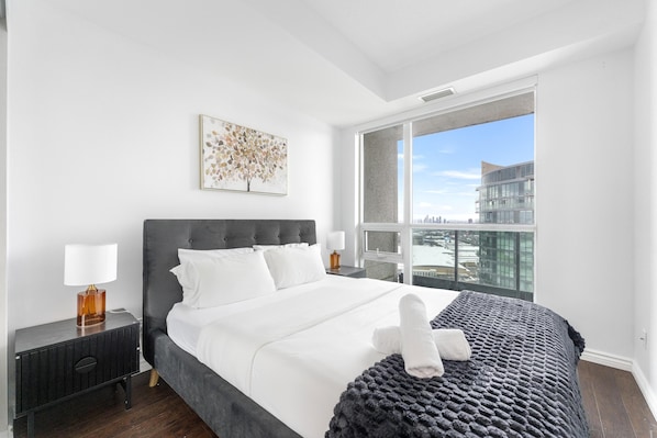 Un dormitorio moderno con una cama grande, una mesita de noche, una lámpara, una ventana con vistas a la ciudad y una obra de arte enmarcada en la pared.