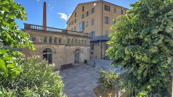 Une cour intérieure avec un bâtiment doté de fenêtres en arc de cercle et d’une cheminée, le tout entouré de verdure.