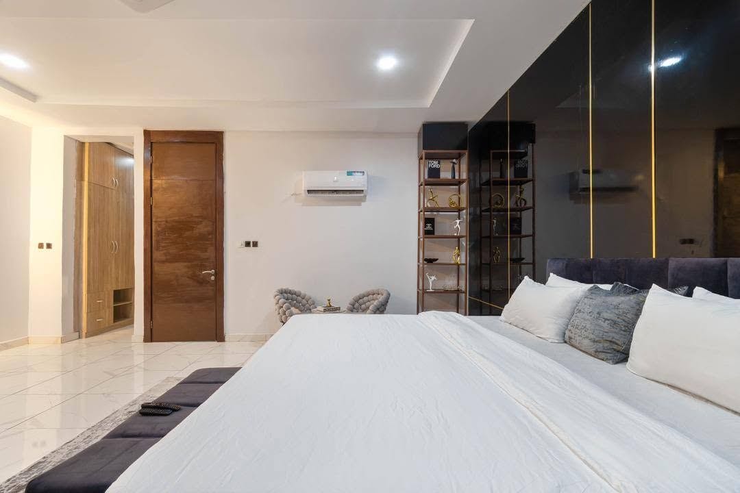 A modern bedroom with a large bed, a wardrobe, a small seating area, and a bookshelf.