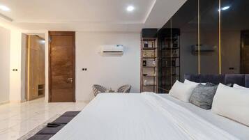 A modern bedroom with a large bed, a wardrobe, a small seating area, and a bookshelf.