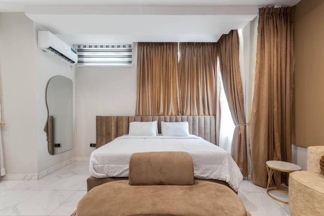 A hotel room with a bed, a headboard, a mirror, a window with curtains, and a small round table.
