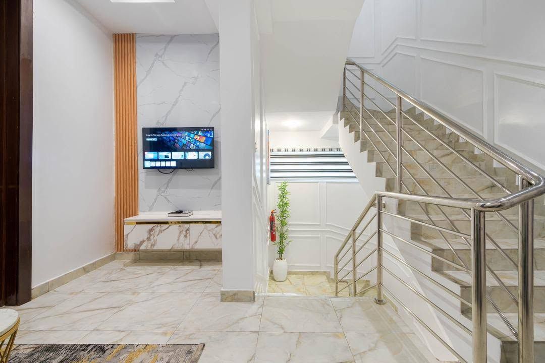 A modern hotel lobby with a marble floor, a flat-screen TV mounted on the wall, a staircase with metal railings, and a potted plant.