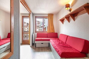 A compact room with a red sofa, a wooden bench, and a small table. A window with curtains is visible, and a wall-mounted shelf is present. - Alpine Ski Apartment in Tignes for 4 (Tignes)