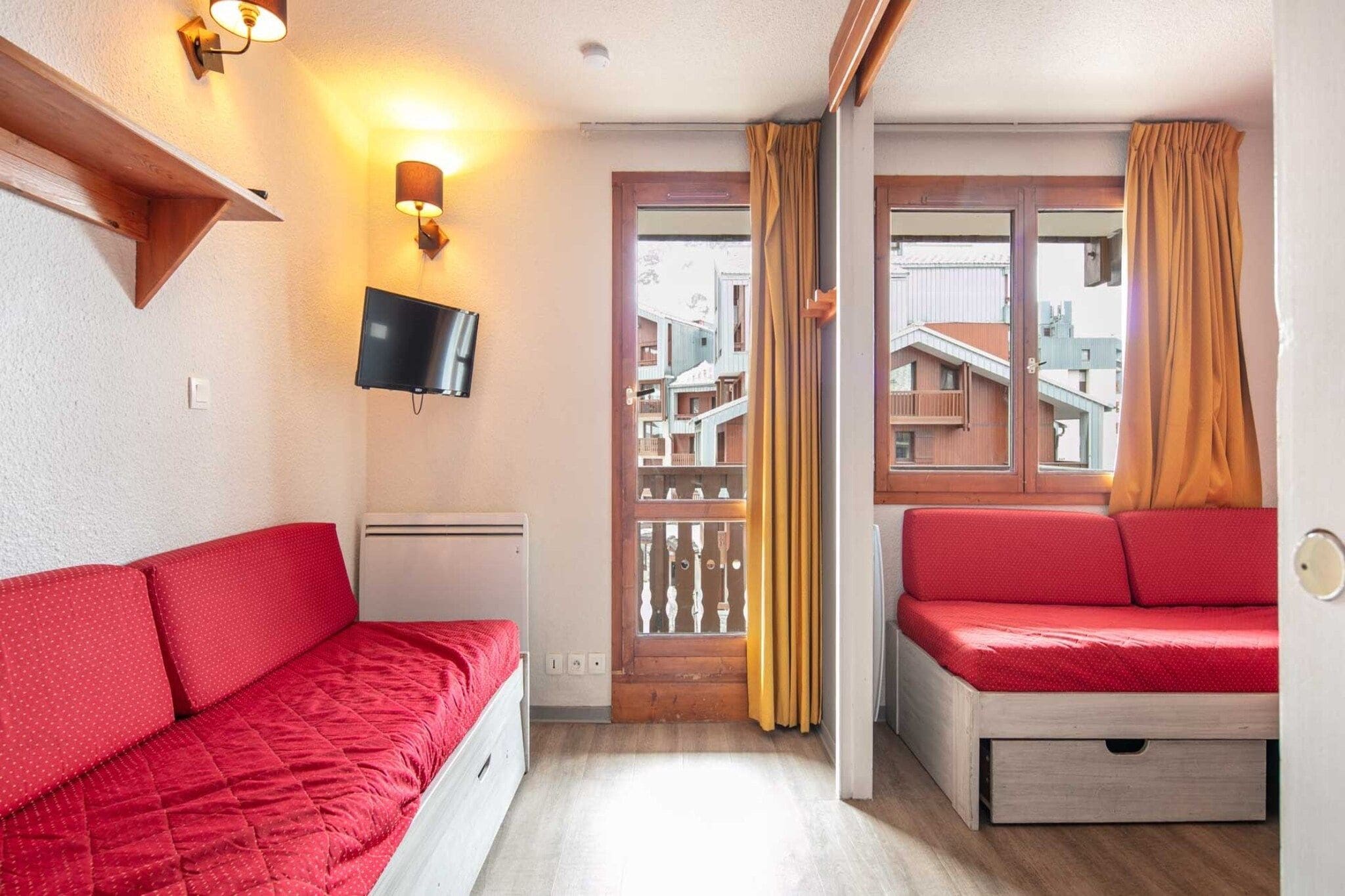 A room with a red sofa, a television mounted on the wall, and a window with curtains.