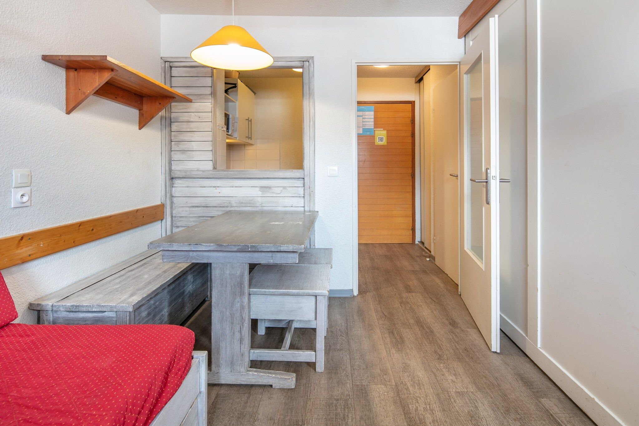 A room with a wooden bench, a small table, a red bed, and a hanging light fixture.