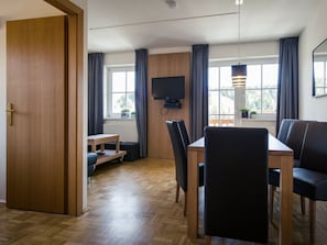 Un comedor moderno con una mesa y sillas de madera, un televisor montado en la pared y ventanas grandes con cortinas.