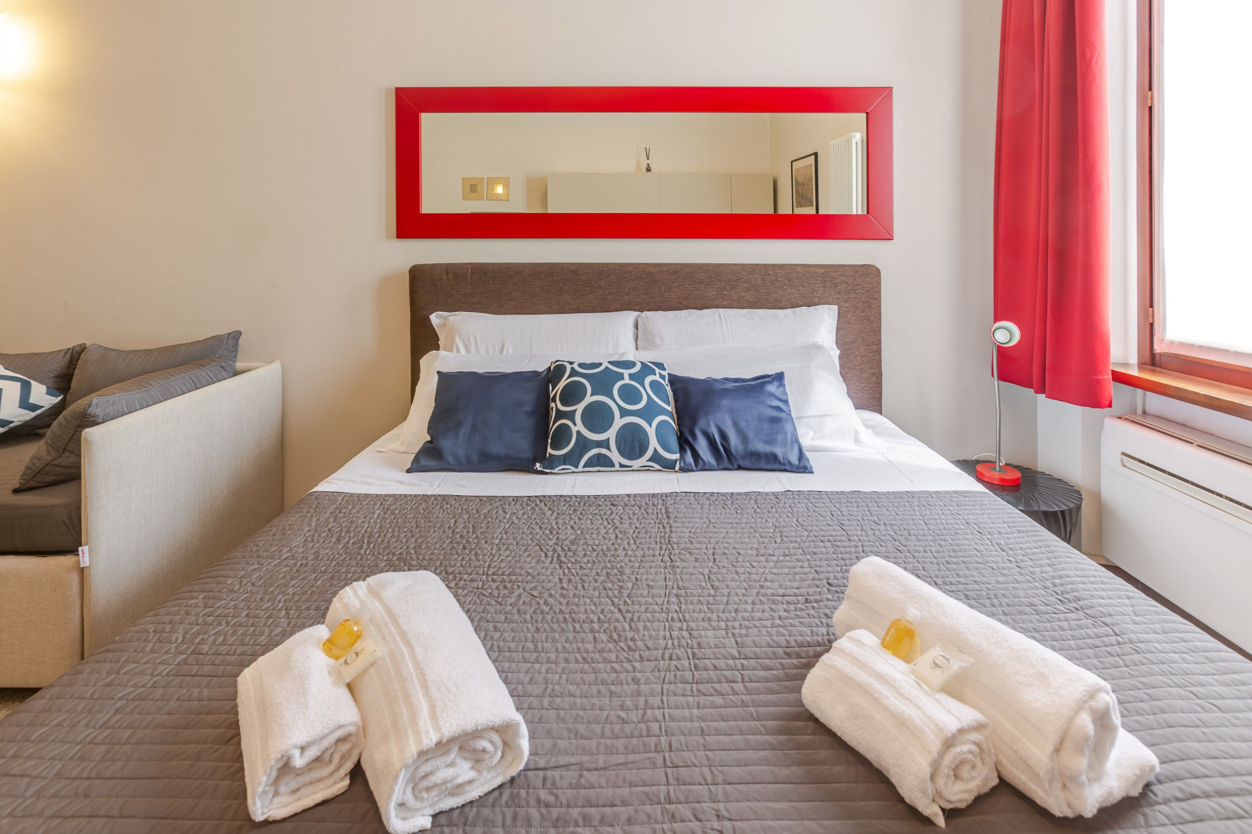 Habitación de hotel con cama, almohadas, un espejo con marco rojo, una mesita de noche, una lámpara y una ventana con cortinas.