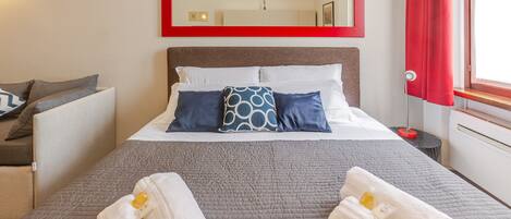 Habitación de hotel con cama, almohadas, un espejo con marco rojo, una mesita de noche, una lámpara y una ventana con cortinas.