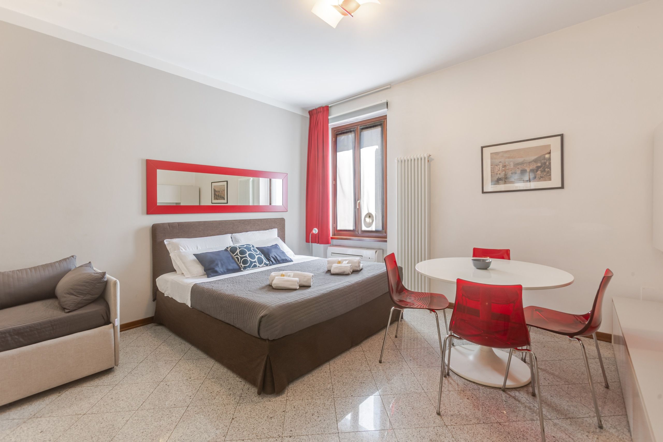 Una habitación de hotel con cama, sofá, mesa de comedor con sillas, espejo, ventana con cortinas y radiador.