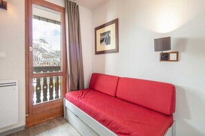A room with a red sofa, a window with a view of buildings and mountains, a framed picture on the wall, and a wall-mounted light fixture. - Cozy Hideaway with Mountain Vistas (Tignes)