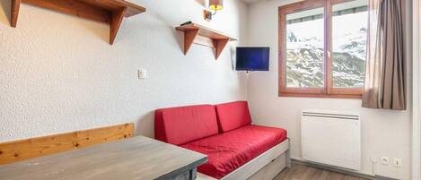 A room with a wooden bench, a red sofa, a television mounted on the wall, and a window with curtains.