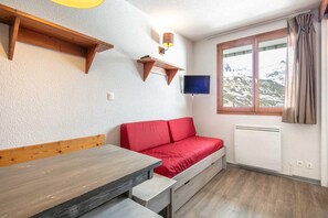 Une pièce avec un banc en bois, un canapé rouge, un téléviseur fixé au mur et une fenêtre avec des rideaux.