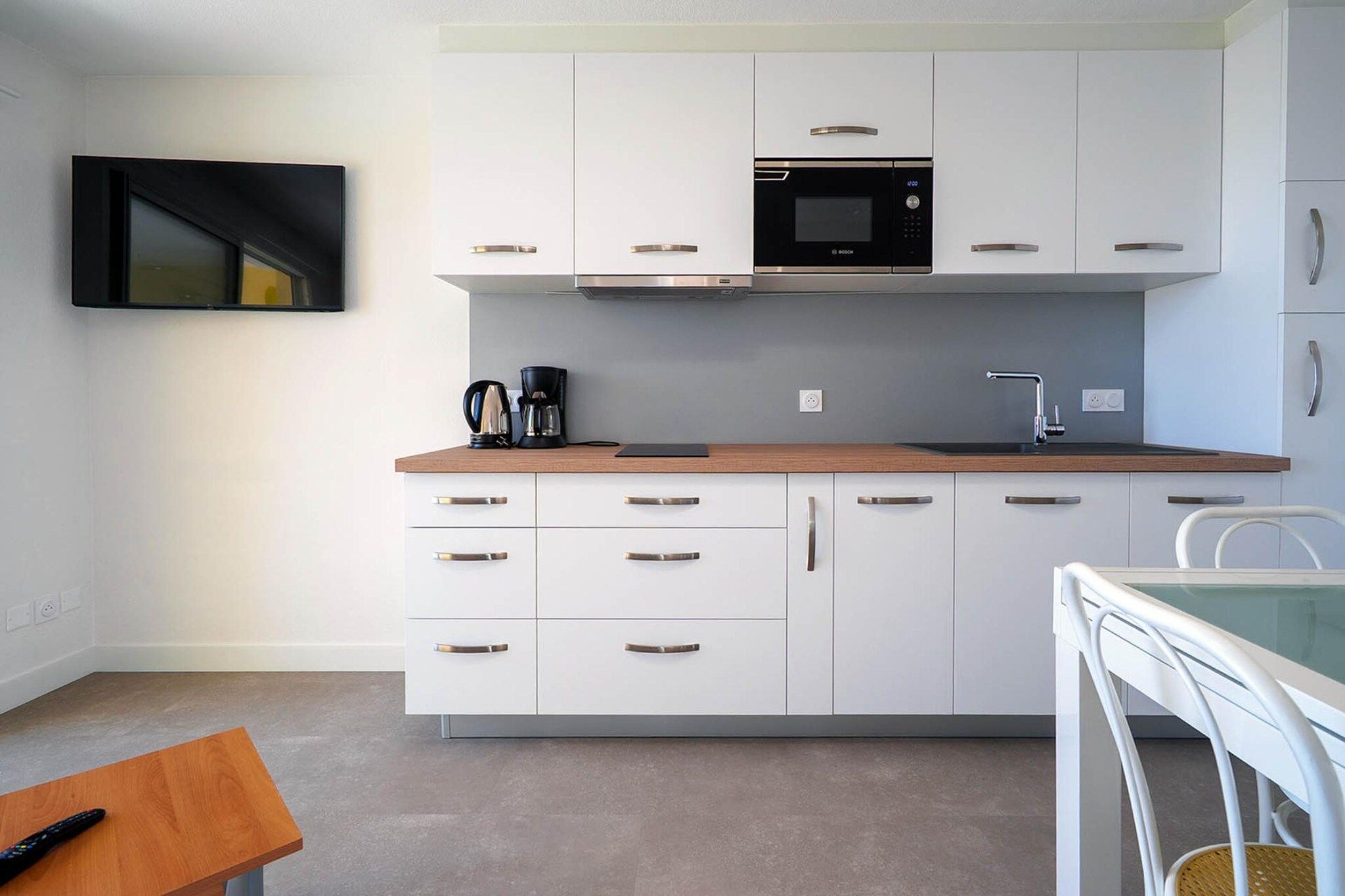 A compact kitchen with white cabinets, a microwave, a coffee maker, and a dining area with a white table and chair.
