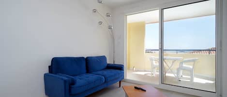 Een kamer met een blauwe bank, een klein houten tafeltje en een glazen deur die leidt naar een buitenruimte met een tafel en stoelen.
