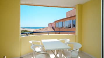 A balcony with a white table and chairs, a view of the beach, and a building with a tiled roof.